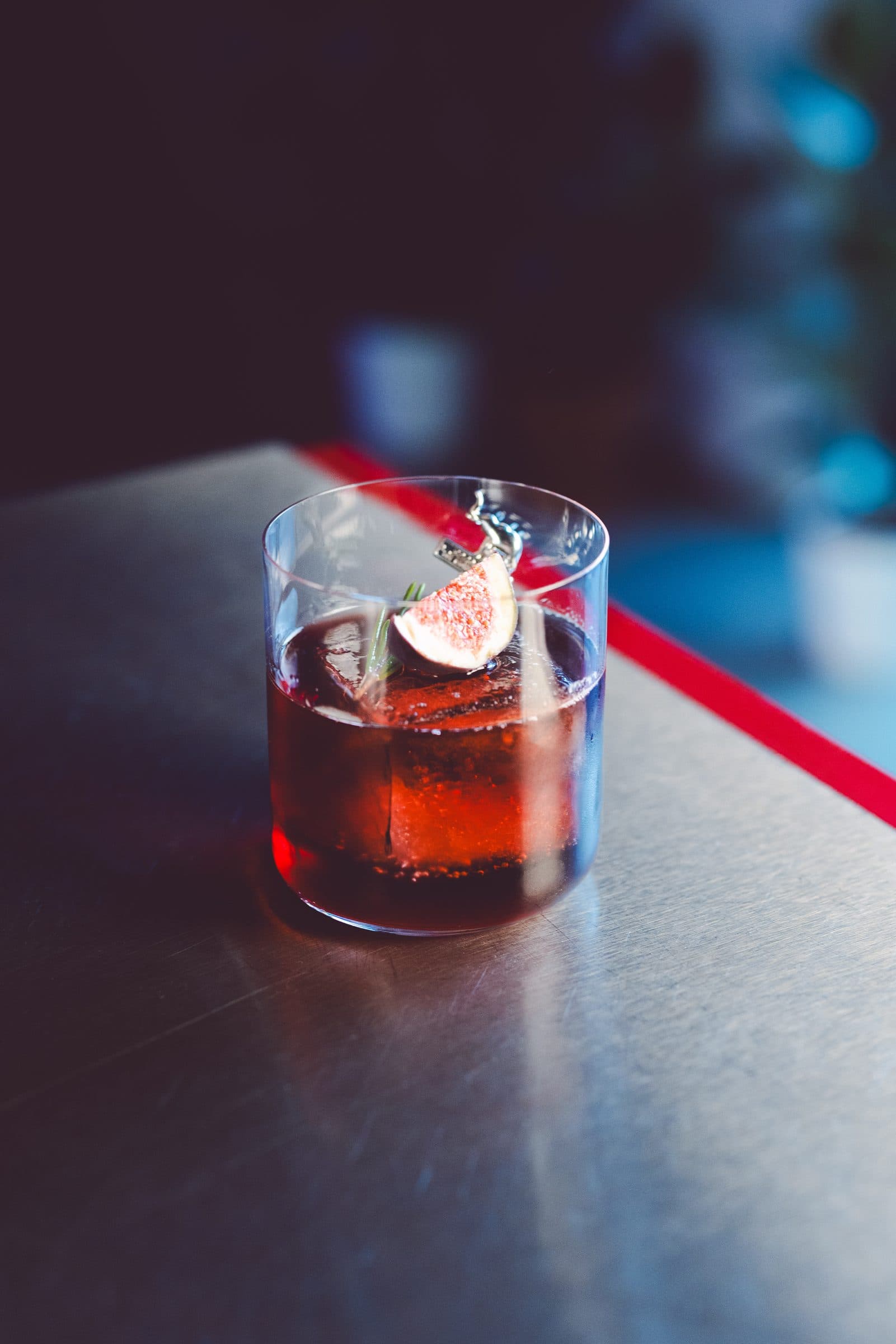 Roter Cocktail im Tumbler mit Feigenspalte und Kräuterzweig auf der Metalltheke des ROMI Studio, dahinter blaues Licht