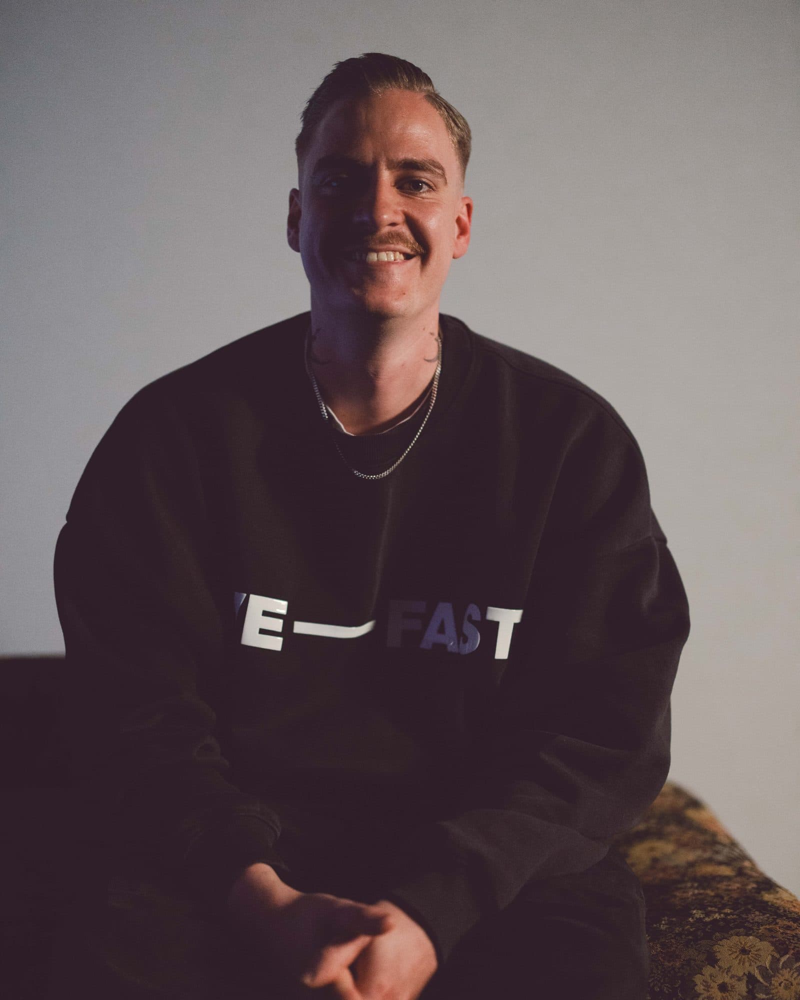 Marvin, Head Bartender von ROMI, sitzt lachend im schwarzen Sweatshirt vor einer hellen Wand